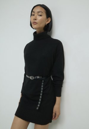 Schwarzes Rollkragenkleid aus weichem Strickmaterial mit geripptem Saum, in der Taille mit einem schwarzen Gürtel mit silbernen Ösen verengt.