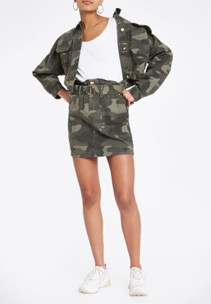 Veste camouflage et jupe assortie dans des nuances de vert. La veste comporte des boutons et des poches ; la jupe a une taille ceinturée et une coupe ajustée. T-shirt blanc et baskets.