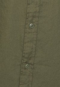 Tkanina koszuli w kolorze oliwkowej zieleni o gładkiej fakturze, z dwoma guzikami w kontrastowych kolorach oraz widocznym przeszyciem wzdłuż listwy z guzikami.