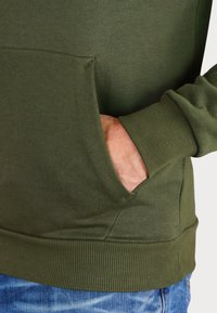 Felpa verde oliva con polsini e orlo a costine, dotata di una tasca frontale. Il materiale è liscio con una leggera texture. Vestibilità casual.