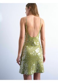 Femme portant une robe courte verte sans manches et dos nu, couverte de sequins réfléchissants, debout devant un fond clair uni.