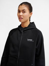 Giovane donna con i capelli raccolti che indossa una felpa nera con zip e logo "hummel" sul petto, rivolta verso di sé su sfondo bianco.
