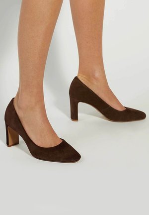 Benen met donkerbruine suède pumps met een middelhoog hakje en ronde neus, tegen een lichte effen achtergrond.