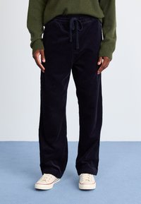 Pantalons en velours côtelé bleu marine avec une taille à cordon de serrage, coupe droite et texture côtelée visible, associés à des baskets beiges.