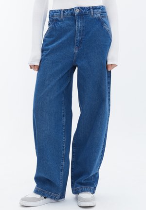 Jeans de mezclilla azul de pierna ancha y cintura alta, con cierre de botón, bolsillos laterales y costuras sutiles. Combinados con una camiseta blanca de manga larga y zapatillas.