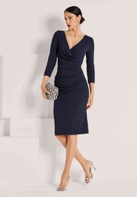Marineblaues Kleid mit Wickeldesign, Drei-Viertel-Ärmeln, knielang, kombiniert mit silbernen Riemchen-High Heels und einer strukturierten grauen Clutch.