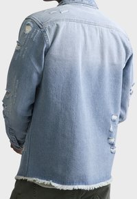 Chaqueta de mezclilla azul claro con detalles desgastados, dobladillo deshilachado y corte holgado. Incluye puños abotonados y costuras de hombros con marcas de uso.