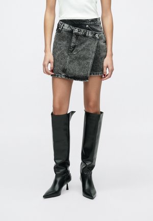 Kobieta nosi czarną asymetryczną mini spódniczkę z denimu z efektem acid-wash, biały top oraz czarne, skórzane kozaki za kolano z szpiczastymi noskami i niskimi obcasami.