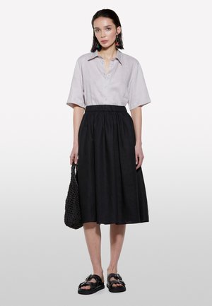 Camicia beige chiaro a maniche corte con bottoni, gonna nera lunga fino alla caviglia, sandali neri con fibbie oversize e borsa nera intrecciata.