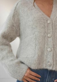 Light gray fuzzy cardigan with a V-neck, featuring four square rhinestone buttons and a front pocket, ribbed cuffs, and soft texture.