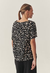 Fekete rövid ujjú felső különböző méretű pöttyös mintával, kerek nyakkivágással és laza szabással, mely sima tapintású anyagból készült.