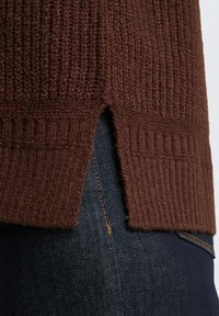 Bruine geribde trui met een split aan de onderrand, gecombineerd met donkere spijkerbroeken. Textuur van gebreide stof en duidelijke stiksels zijn zichtbaar.