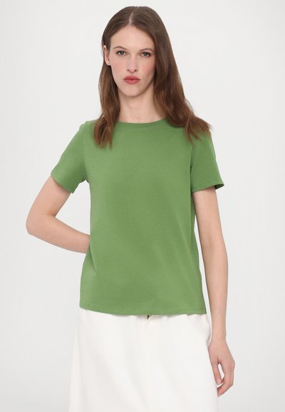 Πράσινο βαμβακερό μπλουζάκι με στρογγυλή λαιμόκοψη και κοντά μανίκια, με χαλαρή εφαρμογή και λεία υφή. Συνδυασμένο με φούστα ανοιχτού χρώματος.