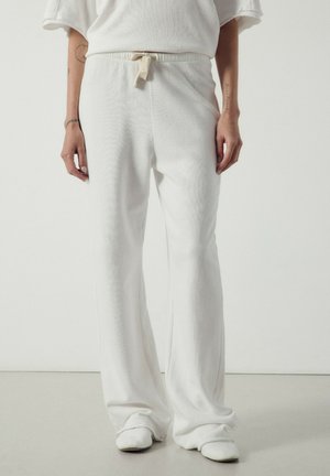 Personne portant un pantalon de jogging blanc ample avec une taille à cordon, des chaussures blanches et un haut blanc sur un fond clair uni.