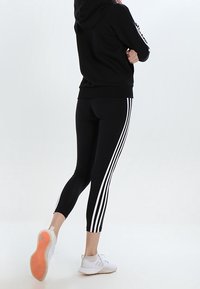 Leggings noirs avec trois bandes blanches sur les côtés, associés à un sweat à capuche noir. Le modèle porte des chaussures de sport blanches. Texture lisse.