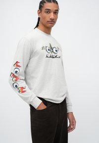 Maglietta grigia a maniche lunghe con una grafica cartoon sul davanti e disegni colorati di occhi sulle maniche. Indossata con pantaloni in corduroy scuri.