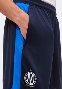 Χέρι στην τσέπη μαύρων αθλητικών σορτς με μπλε πλαϊνό πάνελ και λευκό κυκλικό λογότυπο με αστέρι και κείμενο "Droit Au But" από κάτω.