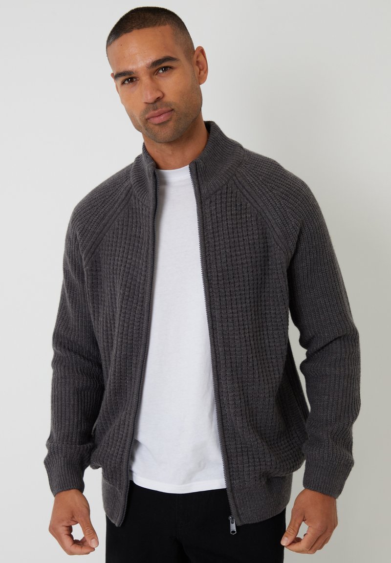 Grauer Reißverschluss-Pullover mit strukturiertem Strickmuster, hohem Kragen und gerippten Manschetten. Über einem weißen Shirt getragen, mit einem glatten Reißverschluss.
