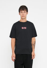 Svart bomull T-skjorte med korte ermer og rund hals. Har et rosa AC/DC-trykk på brystet. Enkelt design, avslappet passform.