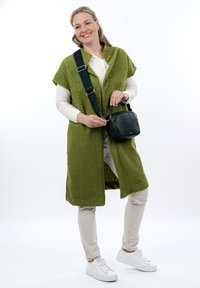 Gilet in pile verde a maniche corte, abbinato a una maglietta bianca a maniche lunghe, pantaloni beige e sneakers bianche. Borsa a tracolla nera compatta.