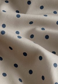 Hellbeige Stoff mit strukturierter Oberfläche, der gleichmäßig verteilte dunkelblaue Punkte in verschiedenen Größen aufweist.