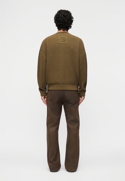 Άνδρας με σγουρά μαλλιά που φοράει καφέ πουλόβερ με υφή και σκούρο καφέ παντελόνι, στέκεται με την πλάτη προς τα εμπρός σε ένα απλό φόντο.