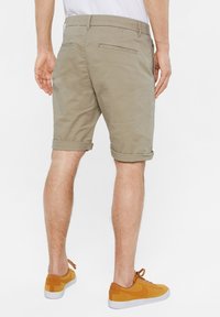 Man wearing rolled-up beige shorts and mustard yellow Nike sneakers, viewed from the back on a white background.