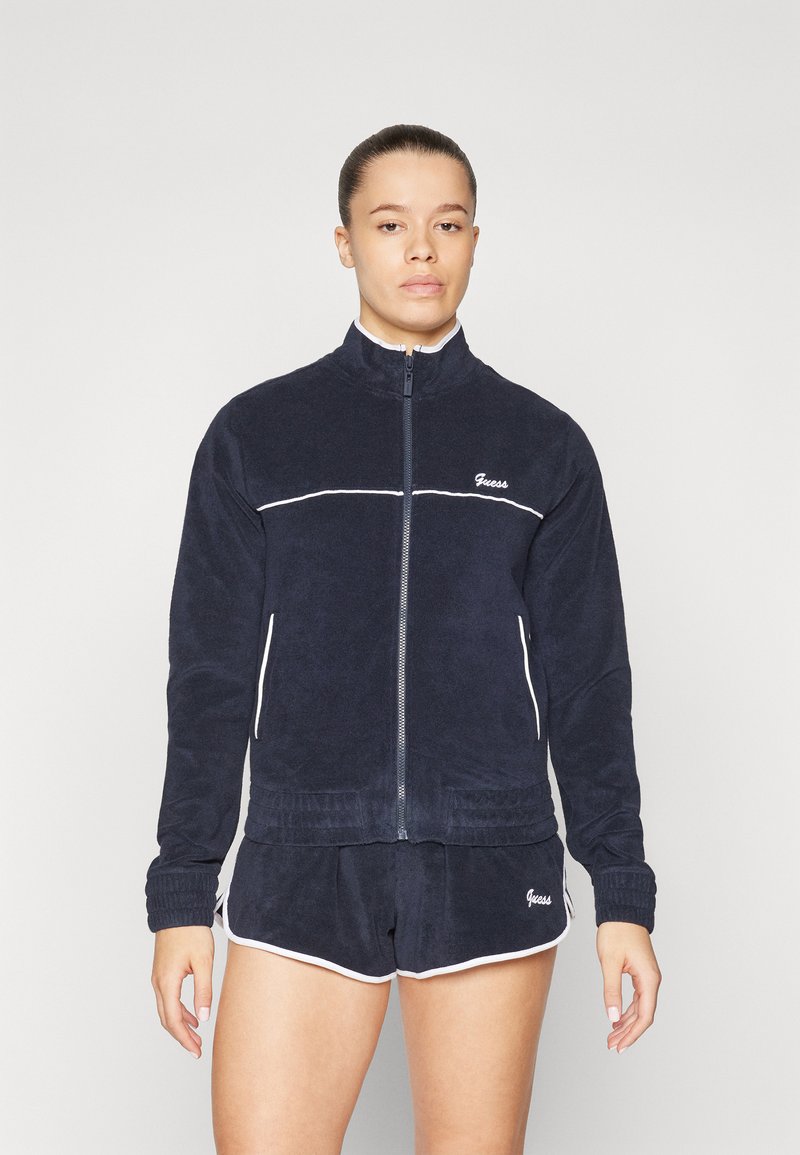 Veste zippée marine avec des surpiqûres blanches, manches longues et poignets côtelés, assortie à un short. Les deux présentent des détails du logo "Guess".