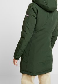 Grüne isolierte Parka mit Kapuze, ausgestattet mit einem Logo-Patch auf dem linken Ärmel und gerippten Bündchen, entworfen für Wärme und Schutz.
