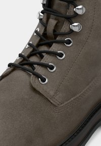Gros plan d'une botte en daim marron avec des lacets noirs passés dans des œillets et crochets métalliques sur une semelle noire.
