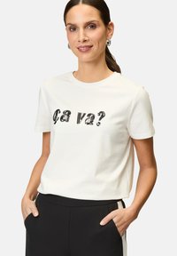 White cotton t-shirt with short sleeves, featuring black sequined text "ça va?" on the front. Worn with black trousers, hands in pockets.