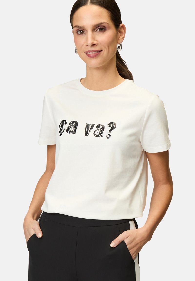 White cotton t-shirt with short sleeves, featuring black sequined text "ça va?" on the front. Worn with black trousers, hands in pockets.
