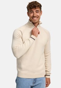 Beżowy sweter z wysokim kołnierzem i zamkiem, z prążkowanymi mankietami i dołem, teksturowanym materiałem oraz małym logo na piersi.