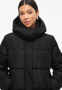 Casaco puffer preto com colarinho alto e dobrado, textura acolchoada e bolsos laterais. Tecido suave com um acabamento liso.