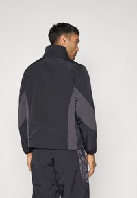 Schwarze Jacke mit hohem Kragen, ausgestattet mit kontrastierenden grauen Seitenpaneelen. Glatte Textur mit subtilen Mustern auf dem Ärmel und Rücken.