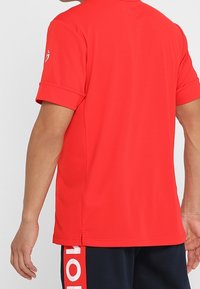 Punainen lyhythihainen poolopaita, joka on valmistettu sileästä kankaasta, ja siinä on klassinen kaulus sekä pieni logo hihassa. Helma on suora.