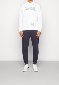 Vit huvtröja med grön grafisk text och illustration, i kombination med mörklila joggers och vita sneakers med gröna detaljer.