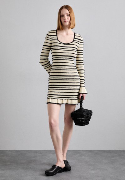 Striped long-sleeve knit dress in cream and black, fitted shape with ruffle hem. Model holds a textured black circular handbag; wearing black clogs.