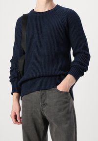 Mörkblå stickad tröja med ribbat mönster och långa ärmar, kombinerad med grå byxor. Har en rund halsringning och sidslitsar.