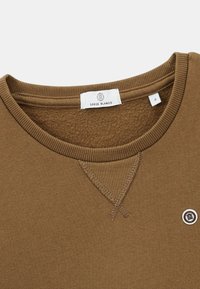 Sweatshirt marron avec un col rond côtelé, texture douce, coutures décoratives et un détail de logo circulaire. L'étiquette indique "Serge Blanco."