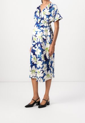 Kobieta w białej sukience o długości do połowy łydki z dużym niebiesko-zielonym kwiatowym wzorem, stojąca na tle białej ściany, w czarnych, niskich butach typu Mary Jane.