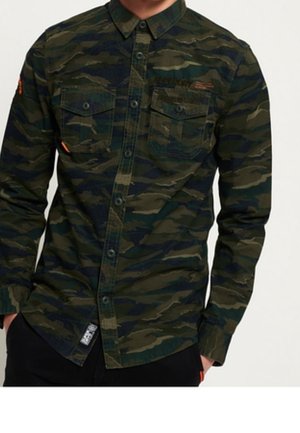 Mand iført en grøn og sort camouflage knap-op skjorte med to brystlommer og sorte bukser, hænderne i lommerne.