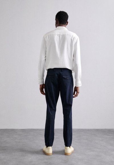 Camisa blanca de manga larga, pantalones azul marino, diseño minimalista. Textura de tela ligera, bolsillos traseros en los pantalones, zapatillas de colores claros.