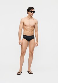 Hemdloser Mann, der schwarze Badehose, schwarze Flip-Flops und schwarze Sonnenbrille trägt und vor einem einfarbigen hellen Hintergrund steht.