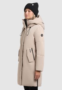 Manteau matelassé beige avec une capuche, poches zippées noires, cordons de serrage réglables et badge logo sur la manche. Bonnet noir côtelé.