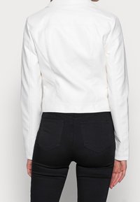 Kobieta w dopasowanej białej kurtce i czarnych dżinsach, pokazana tyłem na jednolitym jasnym tle.