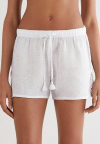 Pantalones cortos de lino blanco con cintura elástica, cierre de cordón y detalles de borlas. Presentan un dobladillo de encaje para un toque adicional.