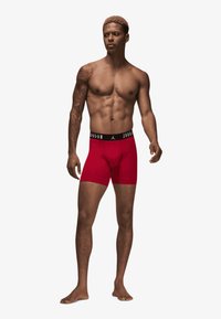 Røde tætsiddende boxer briefs med en sort elastik, der har "JORDAN" branding. Lavet af strækbart materiale, designet til komfort og støtte.