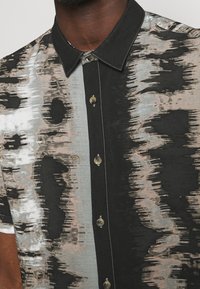 Košile s krátkým rukávem vyrobená z látky s černými a světle šedými abstraktními vzory, s špičatým límcem a béžovými knoflíky.