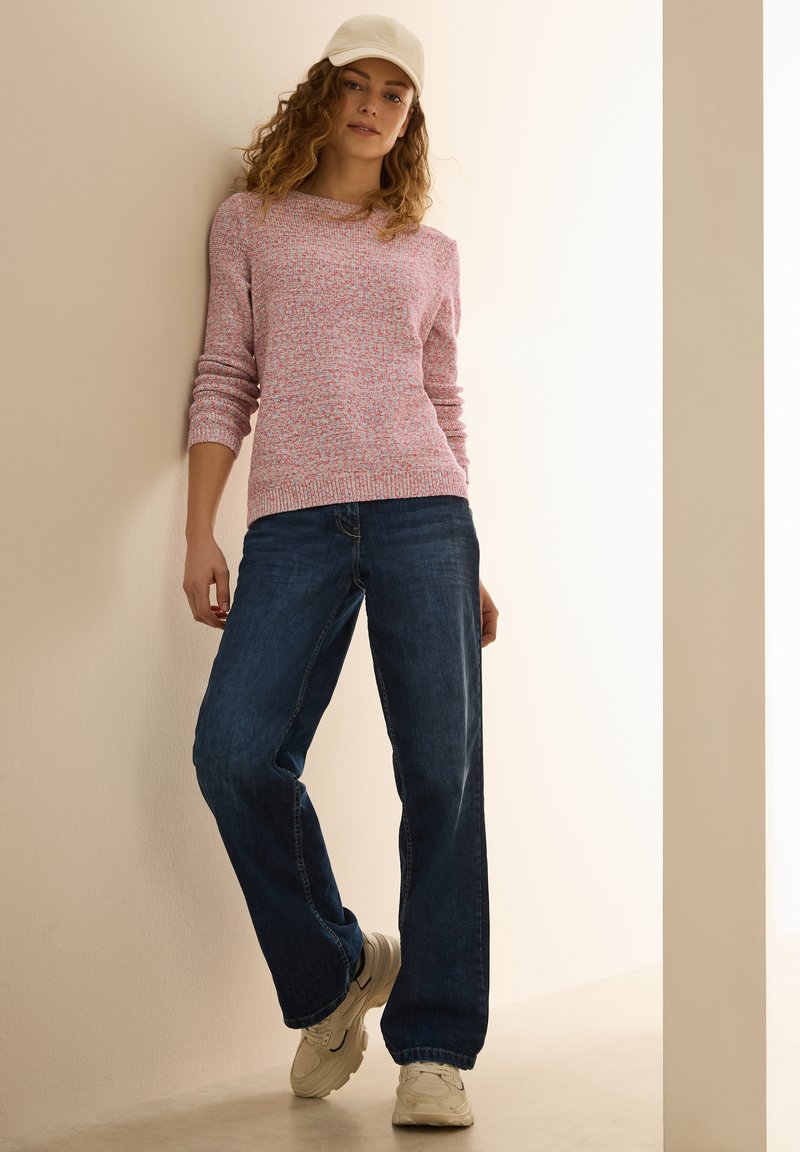 Maglione lavorato a maglia rosa e bianco, con scollatura ampia, abbinato a jeans in denim scuri a gamba larga. Sneakers bianche completano l'outfit.